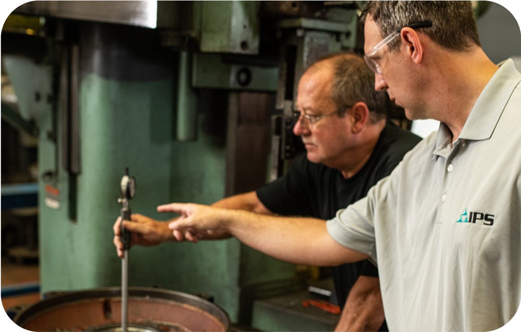 Two men wearing safety glasses in a manufacturing facility, examining a measuring instrument positioned over what appears to be a circular component. One man wears an IPS-branded polo shirt while the other wears black, and they're surrounded by industrial equipment.